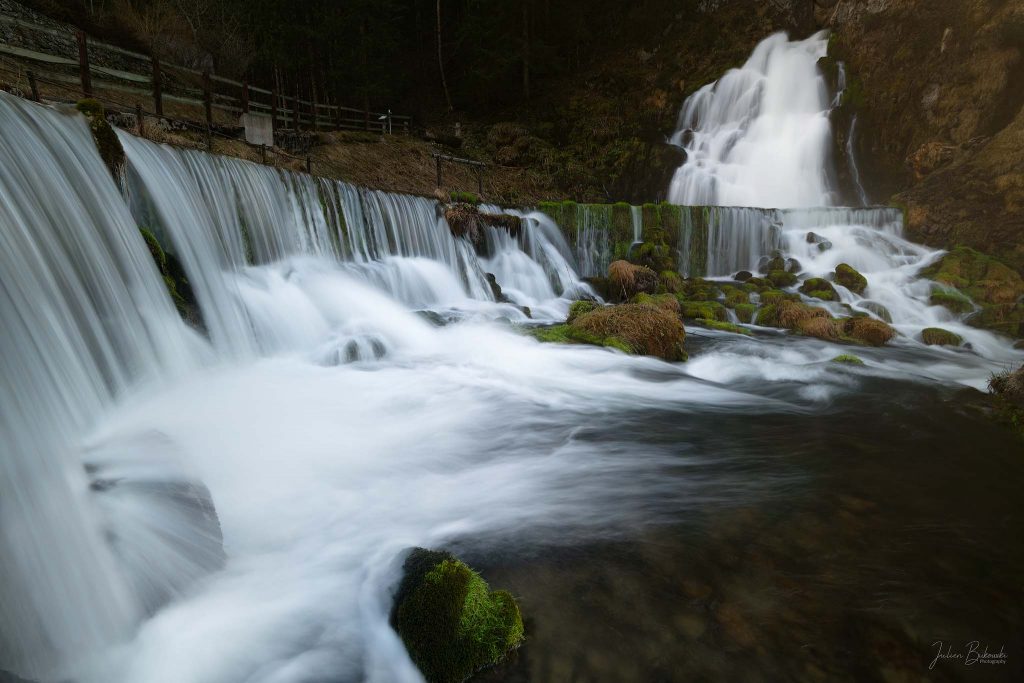 DSC9829 Modifier w Plusieurs rideaux d'eau lissés tombant par-dessus un long seuil en diagonale, blocs couverts de mousse et d'herbe sèche au milieu du courant, cascade principale au fond à droite, barrière en bois et forêt de conifères en haut à gauche, bassin sombre et transparent en bas à droite
